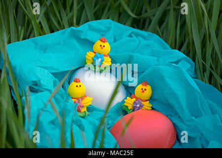 Three toy chickens and three easter eggs on the grass in a blue basket. Composition. Stock Photo