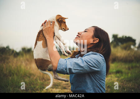 Happy hipster woman smiles and holds a small dog. Little puppy with hipster girls in field sunset background. Vintage tone style. Stock Photo