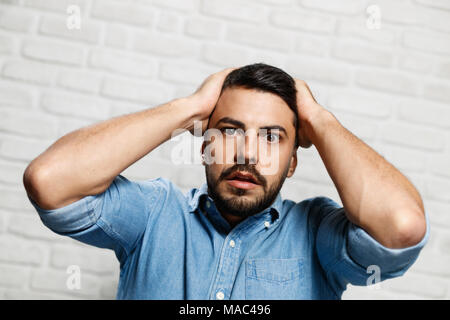 Portrait of upset man against white wall as background. Italian guy getting bad news, touching head with hands. Stock Photo