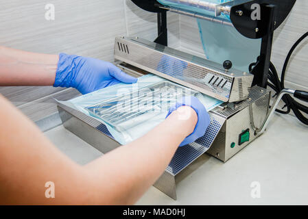Close up dentist's assistant hands in gloves packing dental instrument set for autoclaving in a plastic bag using vacuum packing machine. Sterility and safety care concept. Selective focus. Stock Photo