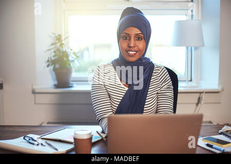 Smiling young Muslim female entrepreneur wearing a hijab sitting in her home office working online with a laptop Stock Photo