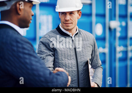 Two smiling engineers wearing hardhats shaking hands together by freight containers on a commercial dock Stock Photo
