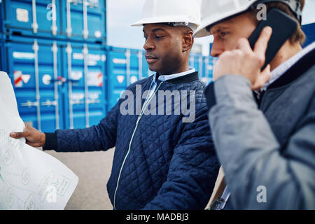 Two engineers holding a building plan and talking on a cellphone while standing by shipping containers in a large commercial shipping yard Stock Photo