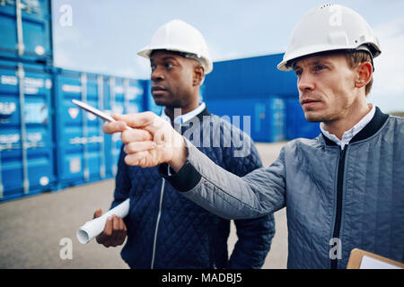 Two engineers wearing hardhats tracking containers together while standing in a large commercial shipping yard Stock Photo