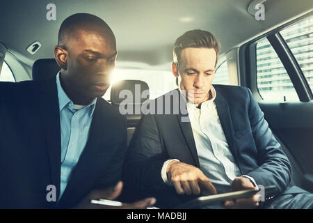 Two diverse businessmen using a digital tablet while sitting together in the backseat of a car driving through the city Stock Photo