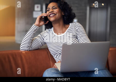 Smiling young African university student sitting on a campus sofa talking on a cellphone and using a laptop Stock Photo