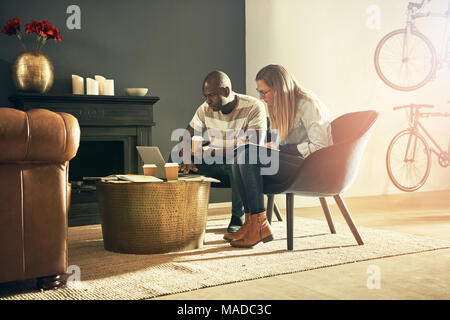 Two diverse young design professionals sitting together in a stylish modern office talking and working online with a laptop Stock Photo