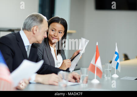 Young businesswoman and her mature colleague discussing structure of report while sitting by desk at conference Stock Photo