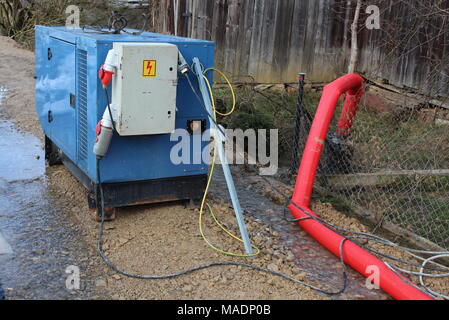 Enclosed industrial power generator providing electrical power to large water pumps with strong red hose pumping water over wire fence during flood Stock Photo