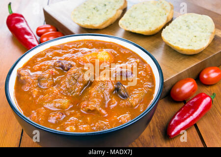 Chilli beef stew in a bowl on a wooden table with garlic breads and raw ingredient of chillies and tomatoes Stock Photo