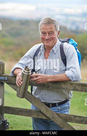 Portrait Of Senior Man On Hike In Countryside Resting By Gate And Having Hot Drink Stock Photo