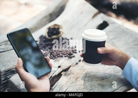 Man hand holding paper cup of coffee with smartphone sitting at wooden table. Stock Photo
