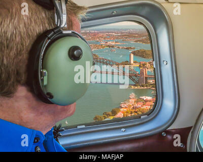 view from a sea plane window flying over Sydney Harbour Australia. A tourist man looks out wearing ear defenders. Stock Photo