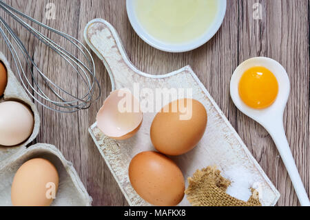 Fresh farm eggs on a wooden rustic background. Separated egg white and yolks, broken egg shells. Whipping eggs with whisk. Preparation of food from ch Stock Photo