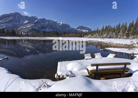 Park Bench covered by snow by tranquil lake, early Springtime in Bow Valley Provincial Park, Alberta Foothills of Canadian Rocky Mountains Stock Photo