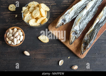 Assorted snacks for beer: sun dried fish, nuts, salted croutons or crackers over wooden background, top view, copy space. Dried smelts. Stock Photo