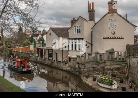 Gnosall, Staffordshire, UK - April 1st 2018 - A canal boat with people enjoy a trip in a narrowboat passing a canal side pub / inn with reflections in Stock Photo