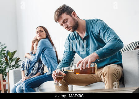 Mother father and son social problems alcoholism sitting on sofa woman protecting her child hugging looking at man drinking whiskey accusingly Stock Photo
