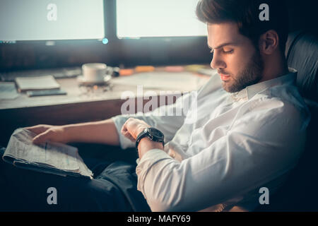 Young bearded man trader at office sitting at table looking at time staying late close-up Stock Photo