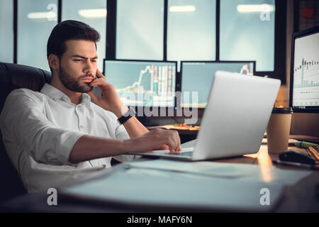 Young bearded man trader at office sitting at table browsing internet on laptop concentrated thoughtful Stock Photo