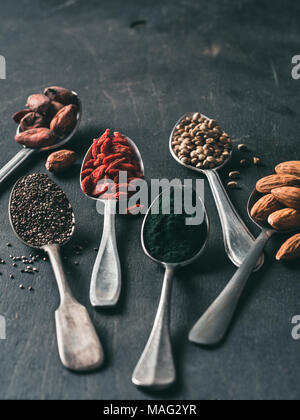 Spoons of various superfoods on black wooden table background. Superfood close up as chia seeds, spirulina, cocoa bean, goji berry, hemp seeds, almond. Copy space for text. Vertical Stock Photo