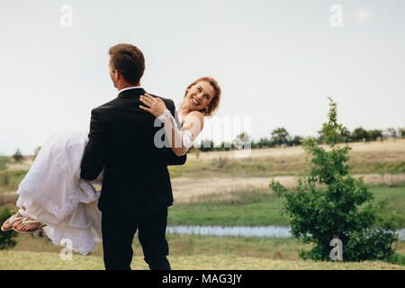 Bridegroom carrying his bride at the park. Newlywed couple having fun outdoors. Stock Photo