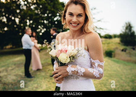 Portrait of beautiful bride holding flower bouquet looking at camera and smiling with guest in background. Bride looking happy at wedding party. Stock Photo