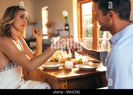 Romantic couple sitting at dining table at home. Man giving a rose to his girlfriend while sitting at dining table. Stock Photo