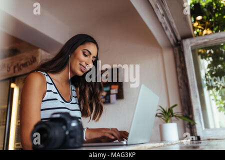 Woman blogger using laptop at home wearing earphones. Woman sitting with a professional camera on the table working on her laptop computer. Stock Photo
