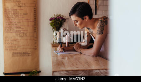 Woman standing at the counter of her cafe noting orders. Restaurant owner making notes standing at the billing counter. Stock Photo