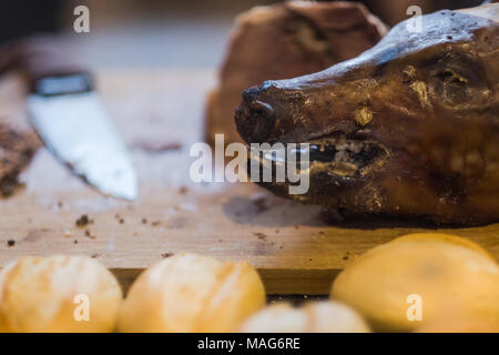 The head of a whole roasted pig resting on a chopping board with the rest of the meat being served as snack in between a bread roll lying in front Stock Photo