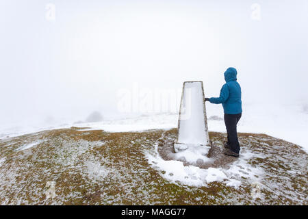 Flintshire, Wales, UK 2nd April 2018, UK Weather: A washout Easter Bank Holiday ends with a Met Officer weather warning for rain and snow for Bank Holiday Monday. A walker resting against a snowy windswept trig point on the highest point of Halkyn Mountain in Flintshire  © DGDImages/Alamy Live News Stock Photo