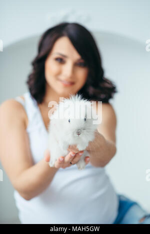 Small lovely white fluffy rabbit in the hands of beautiful smiling pregnant woman at the blurred background. Close-up shot. Stock Photo