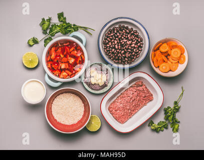 Flat lay of bowls with cooking ingredients for balanced one pan meal with beans, minced meat, rice and various cut vegetables on gray background, top  Stock Photo