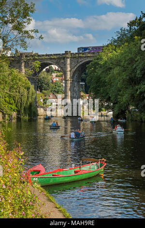 People relaxing & boating in rowing boats on River Nidd under blue summer sky, as train passes over bridge in scenic sunny  Knaresborough, England, UK Stock Photo