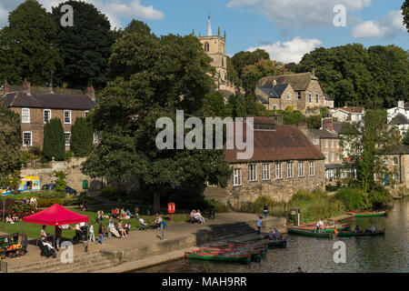 People relaxing by riverside & boating in rowing boats on River Nidd under blue sky - scenic sunny summer view by bridge, Knaresborough, England, UK. Stock Photo