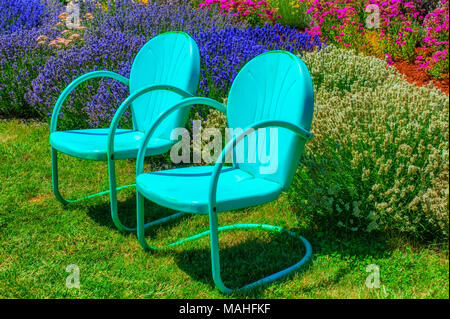 Two blue metal retro lawn chairs invitingly sit on a lawn in a flower garden. Stock Photo