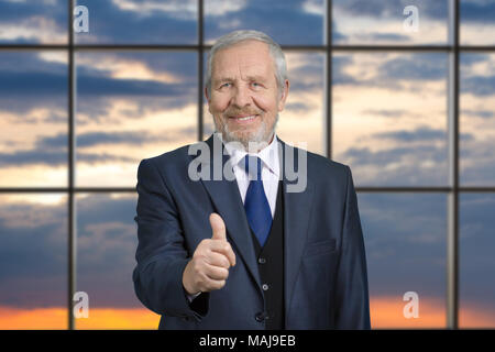Portrait of old TV presenter with thumb up. Senior businessman in suit against windows background with evening sky. Stock Photo