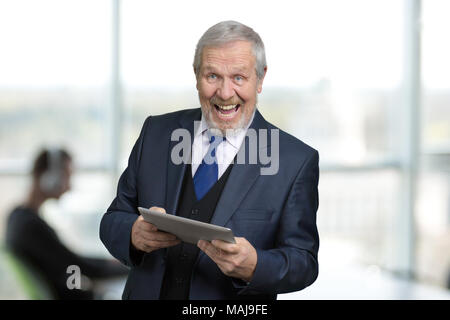 Extremely happy man in suit with tablet. Portrait of rejoicing old manager in office. Bright windows background. Stock Photo