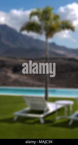 Cropped shot with bokeh effect of residential, hospitality outdoor setup with small palm tree by the pool and white chaise lounge positioned on grass Stock Photo