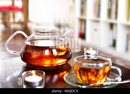 Teatime scene with glass tea pot and cup of tea, old book and hot steam. Idyllic tea time or relaxation scene with cookies and candles. Stock Photo
