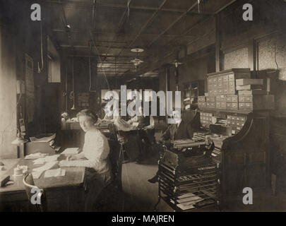 Horizontal photograph of the interior of an office space with men and women sitting at desks and standing. Title: Women and Men Working in Office at Standard Adding Machine Company, 3701 Forest Park Boulevard.  . May 1910. Stock Photo