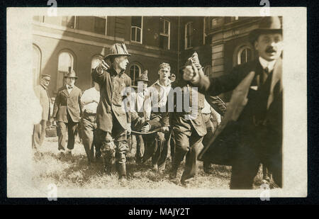 Horizontal photographic postcard showing fireman carrying an occupied stretcher away from a building. Firemen are wearing jackets and hats, and a non-fireman wearing a suit and bowler hat is standing at the far right gesturing to someone off-camera. Title: Firemen of Engine Co. 50 carry casualty away from fire on stretcher.  . circa 1915. Stock Photo