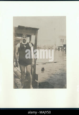 Wet man in a swimsuit standing on a wooden dock beside a small lake with a man swimming. Title: W.E. Dickey of the New York Athletic Club, as the Plunge for Distance champion at the 1904 Olympics.  . 1904. Stock Photo