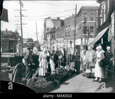 Horizontal, black and white photograph showing shoppers gathered by sidewalk produce outside the Theodore Prieshoff Grocery at 1929 East Grand Avenue, near the Grand Avenue water tower. The produce is arranged on a low platform on the sidewalk, and a small group of men and women is gathered around. Some shoppers are holding baskets full of produce. Several street cars and a horse and wagon can be seen on the left and the Theodore Prieshoff Grocery can be seen on the right. A variety of two and three-story brick buildings are in the background. The print appears to have been made from a broken  Stock Photo