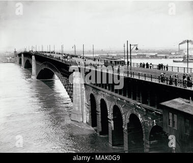 Horizontal, black and white photograph showing the Eads Bridge during high water in 1903. The bridge spans the center of the image. Pedestrians are crossing the bridge and peering over the edge at the Mississippi River. A streetcar travels down the bridge near the center of the image, and several small carriages or wagons are behind it. Title: Pedestrian and trolley traffic on Eads Bridge during 1903 flood.  . 1903.