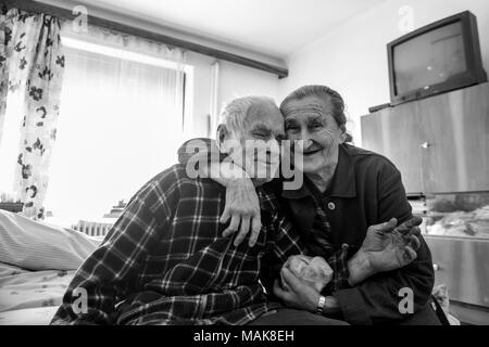 Cute 80 plus year old senior married couple hugging and smiling portrait. Black and white waist up image of happy elderly couple sitting on a bed. Stock Photo