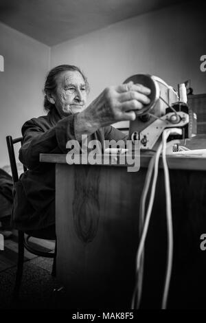 Cute 80 plus year old senior woman using vintage sewing machine. Black and white image of adorable elderly woman sewing clothes in her old family home. Stock Photo