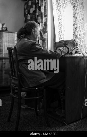 Cute 80 plus year old senior woman using vintage sewing machine. Black and white image of adorable elderly woman sewing clothes in her old family home. Stock Photo