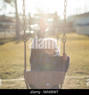 Young boy smiles full of hope and joy reaching for the Spring sunshine white he swings on the swing-set. Stock Photo